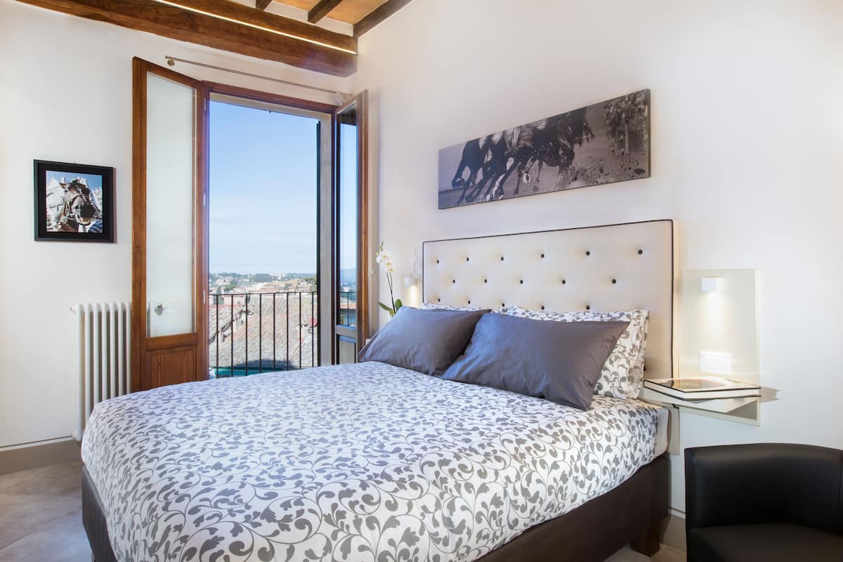 A bedroom with a large bed, a nightstand, a chair, a window with a view, and a picture on the wall.