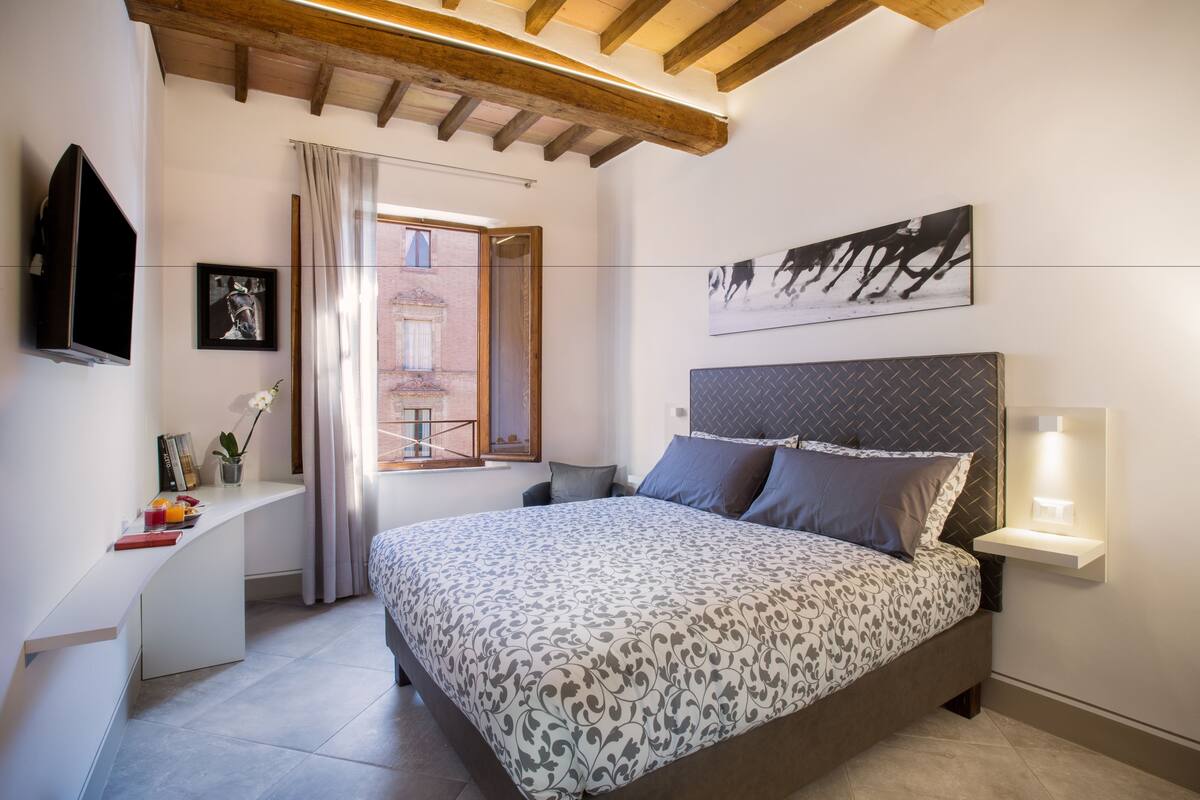 A bedroom with a bed, bedside table, TV, artwork, and a window with curtains.