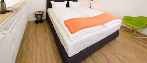 A neatly made bed with white and orange bedding, a green chair, and a radiator on the left.