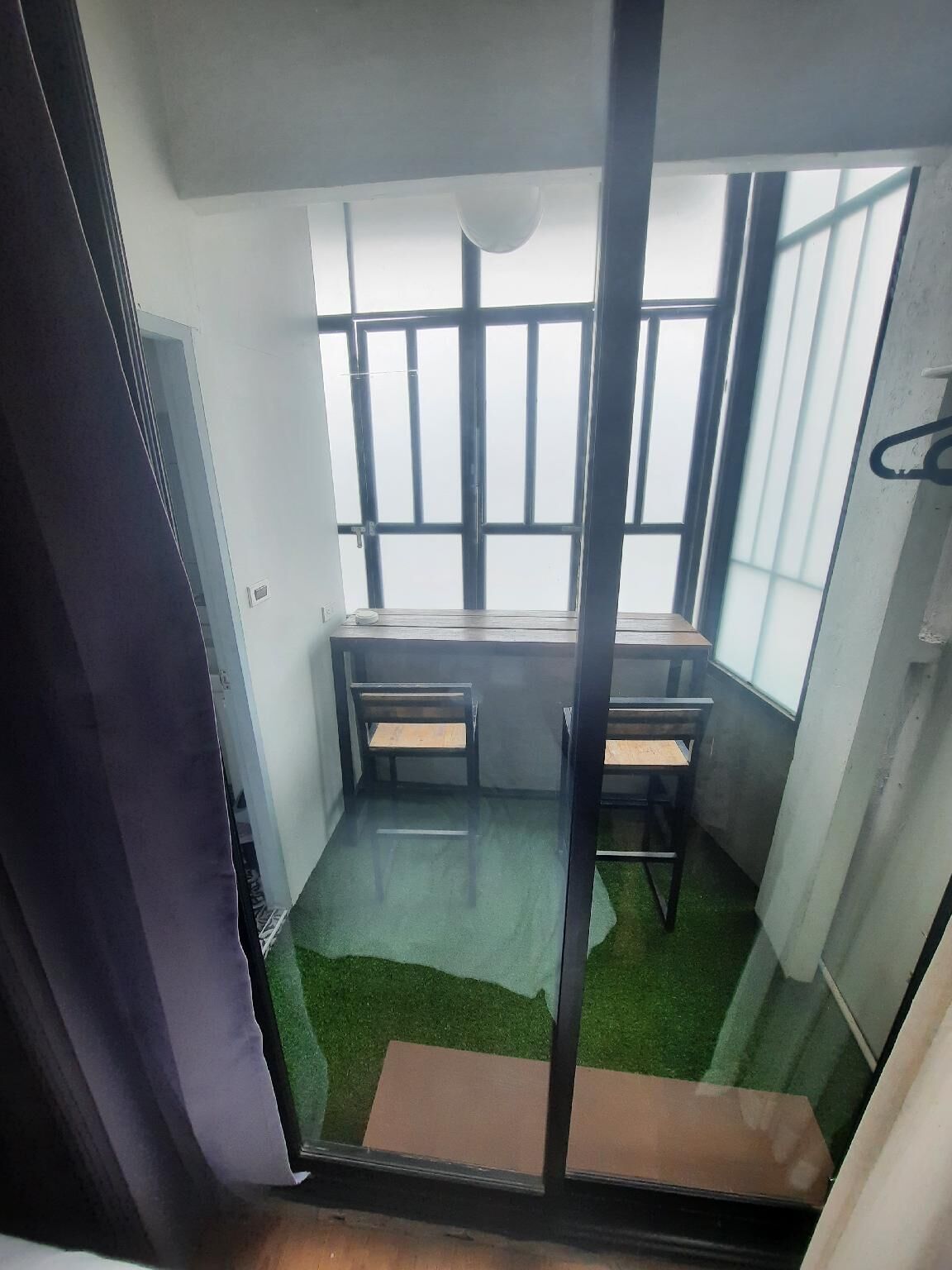 Quarto com escrivaninha, duas cadeiras, janela com grades e piso de grama sintética verde.