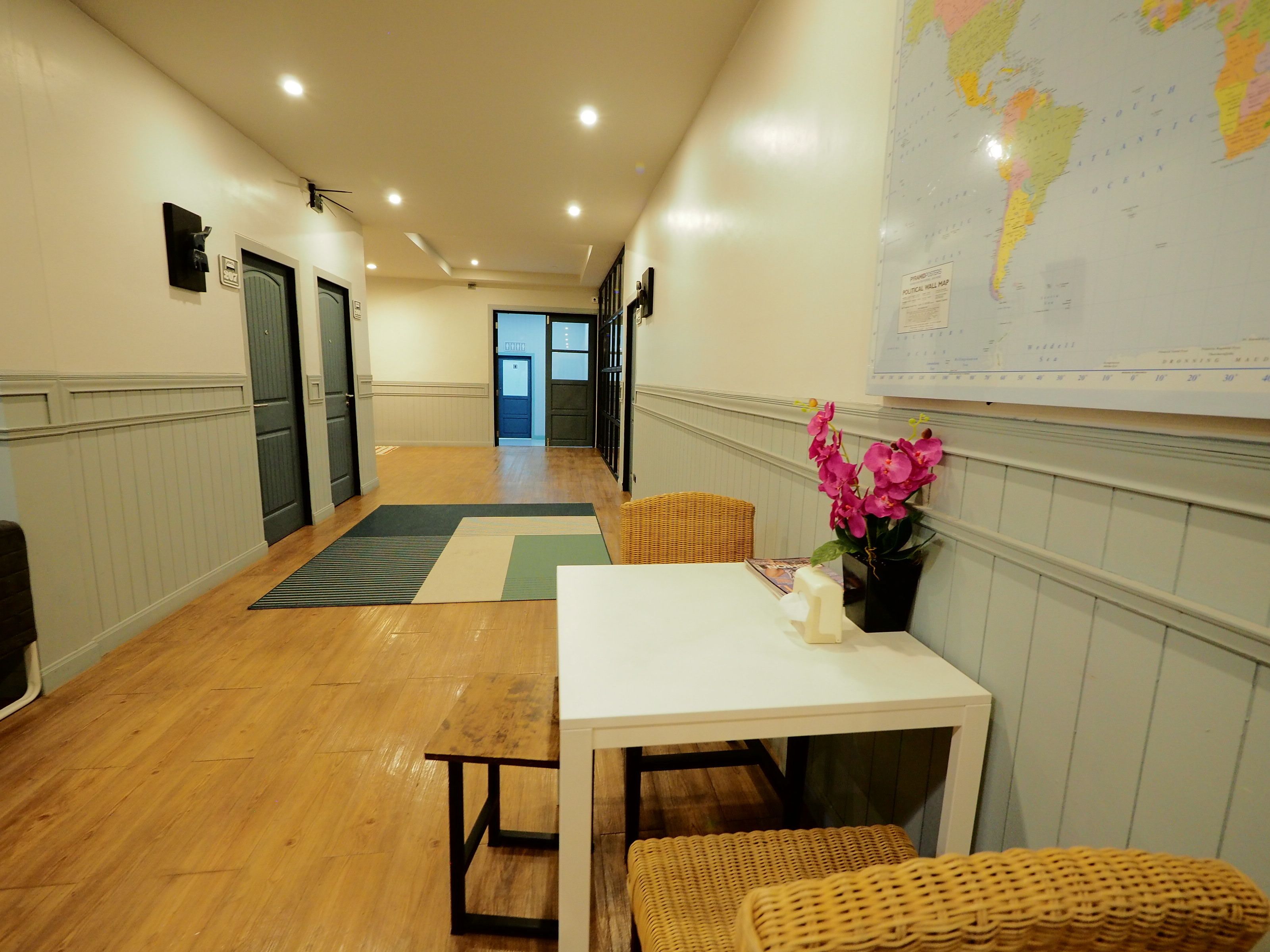 A hotel corridor with a wooden floor, a white table with a vase of flowers, wicker chairs, and a map on the wall.
