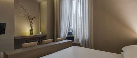 A modern hotel room with a large bed, a seating area with a bench and cushions, a bedside table with a vase, and sheer curtains over a window.