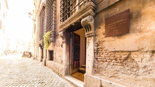Una calle adoquinada con un edificio histórico que presenta una placa con la inscripción "Hotel del Barbiere Palazzo Lazzaroni Roma 1st".