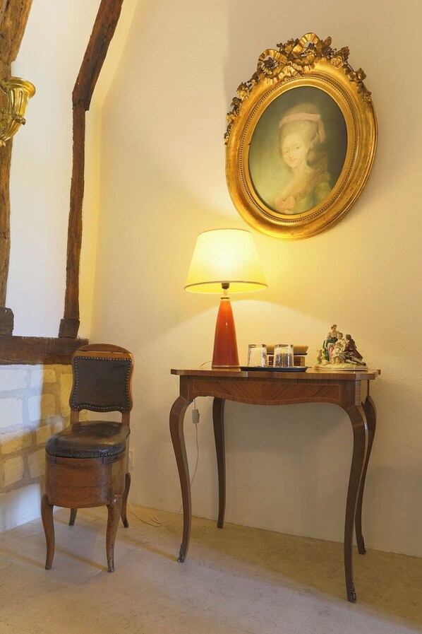 A room with a wooden chair, a table with a lamp, and a framed painting on the wall.