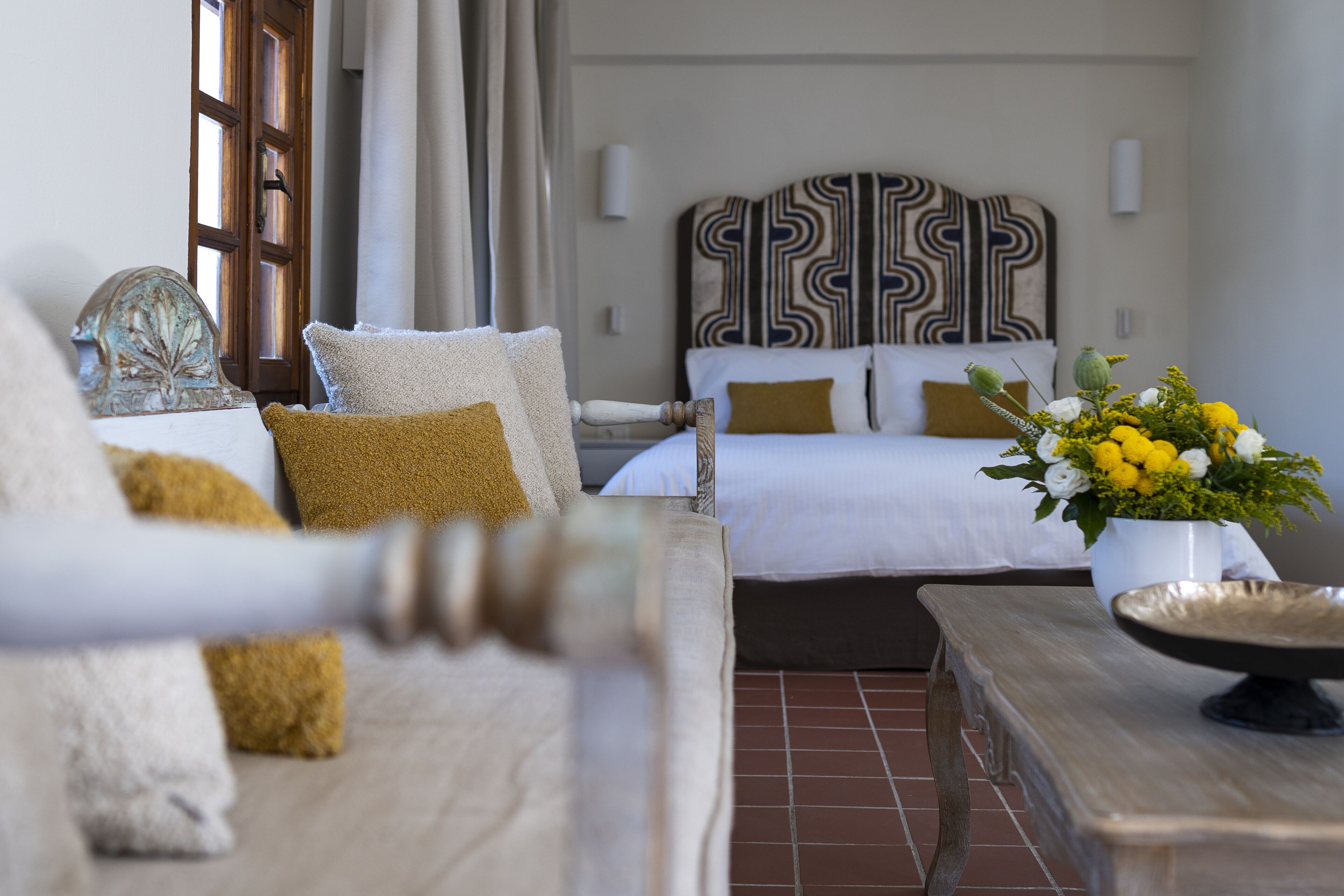 Ein Hotelzimmer mit zwei Betten, einem hölzernen Kopfteil, einem Nachttisch mit Blumenvase und Blick ins Freie durch ein Fenster.