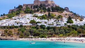 A coastal town with white buildings, a castle on a hill, and a beach with people and boats. - Maris Lindos, Suites and Apartments (Rhodes)
