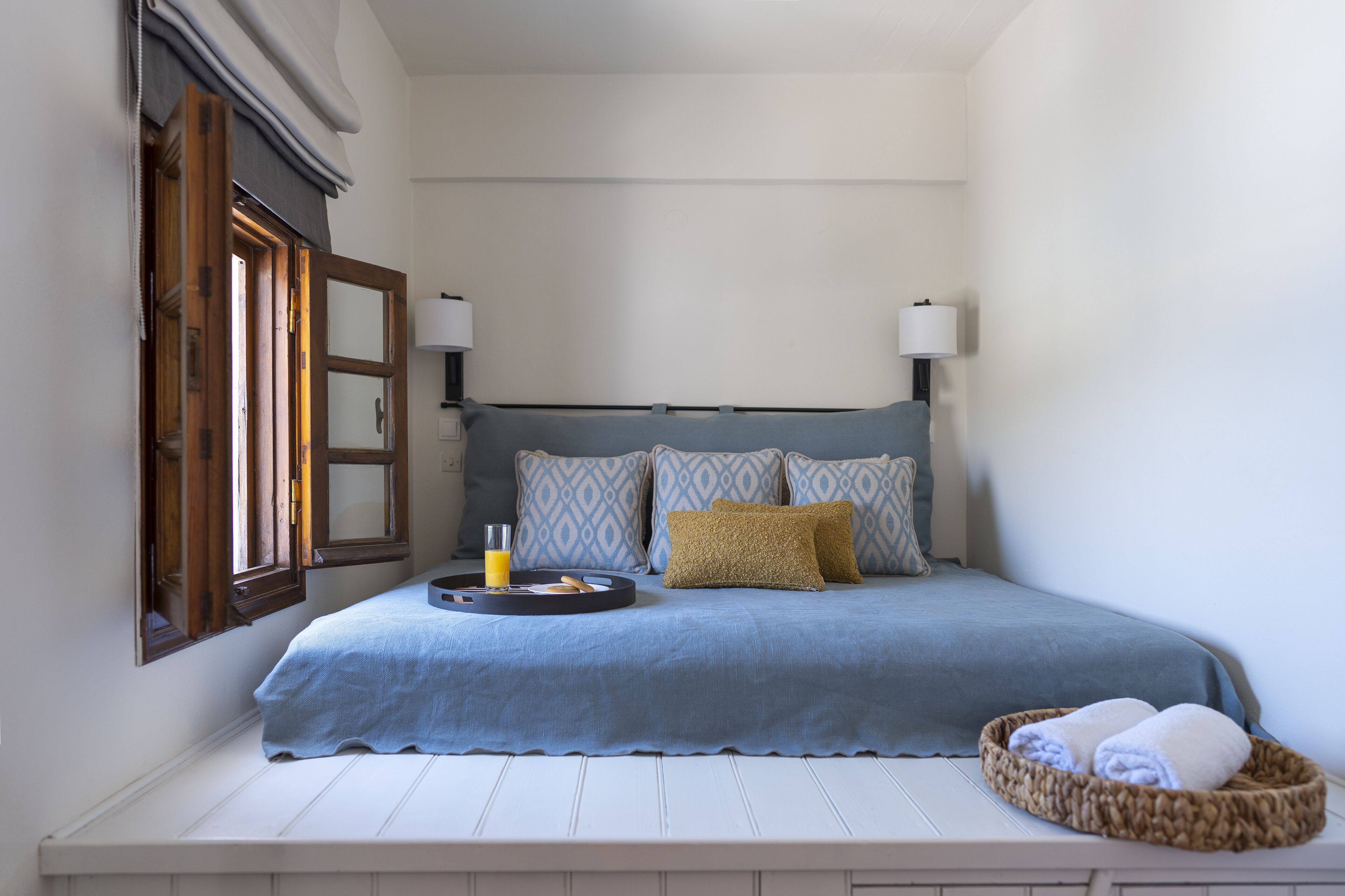 A neatly made bed with a blue bedspread, two pillows, and a bedside table with a tray holding a glass of orange juice and a book.