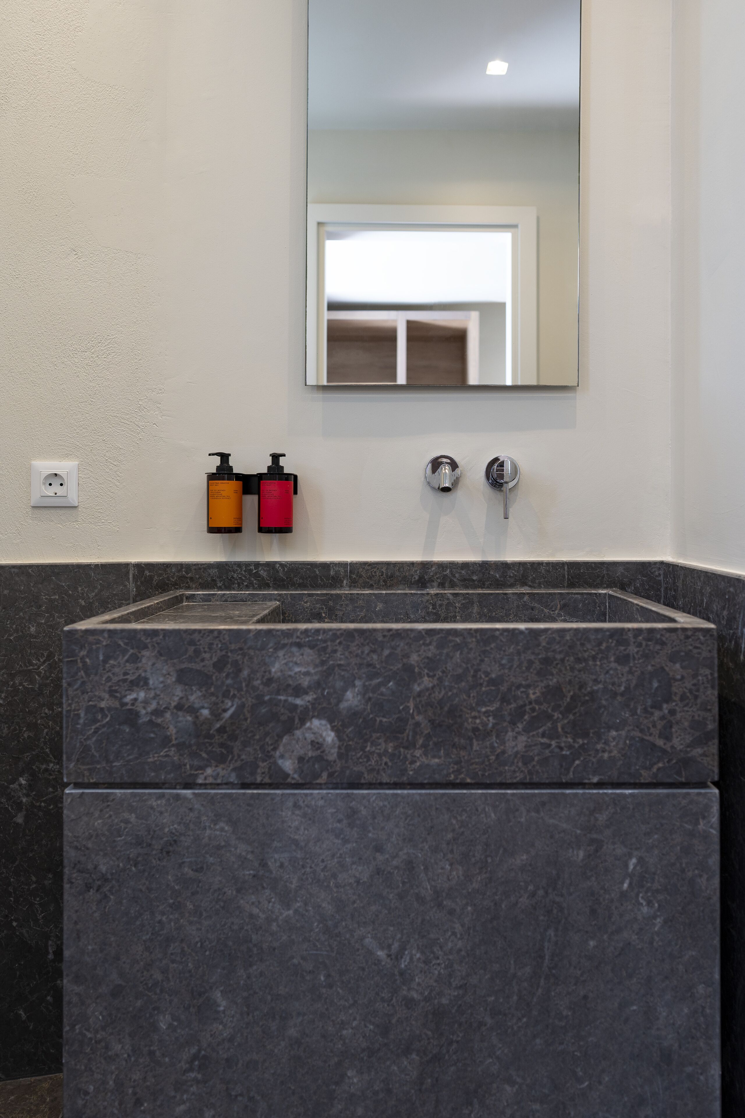 A bathroom with a marble sink, two hand soap dispensers, a mirror, and a wall socket.