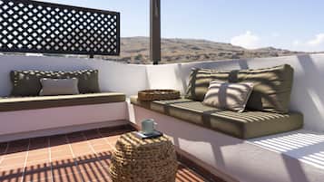 A rooftop terrace with a cushioned seating area, a woven ottoman, and a view of distant hills.