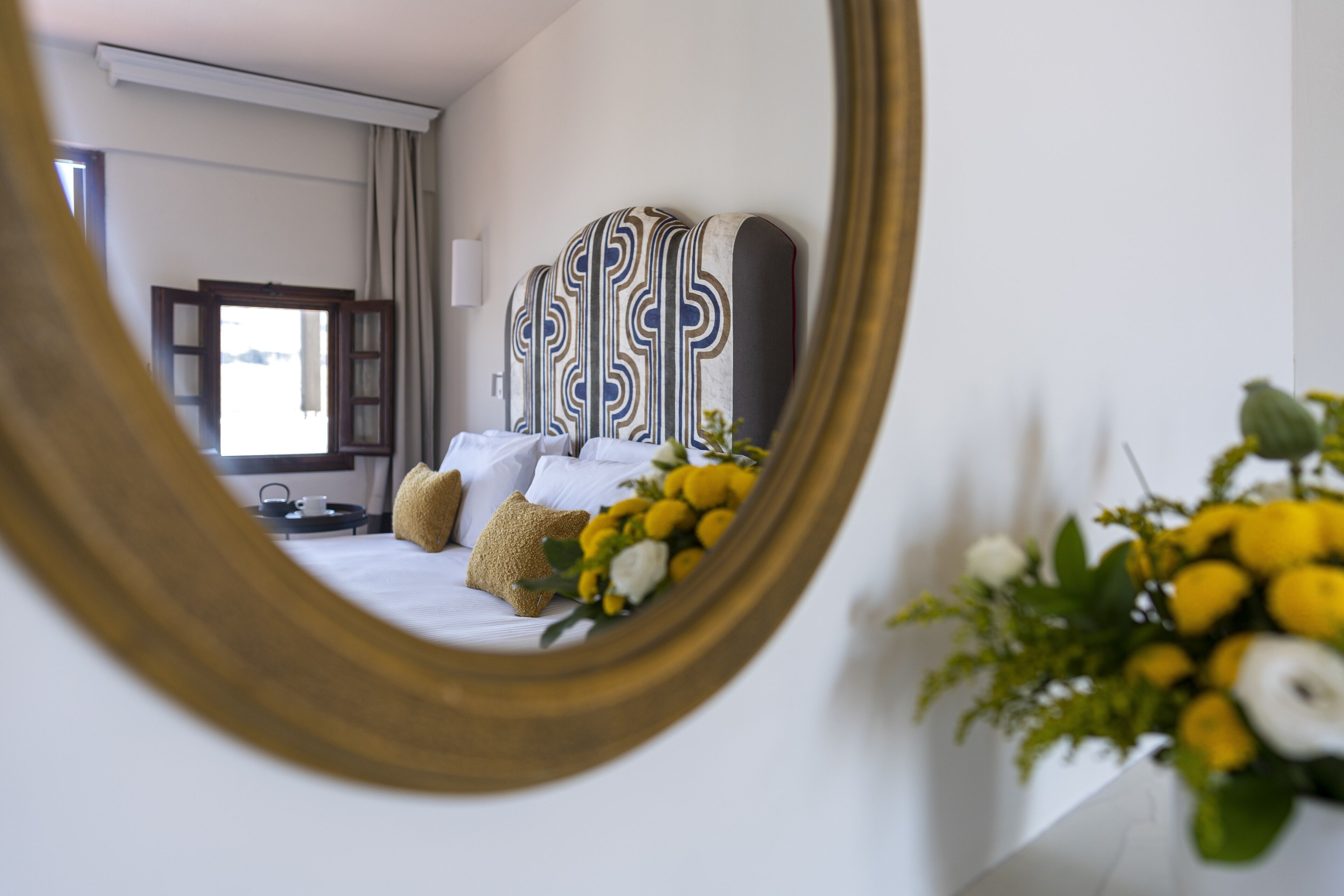 Una habitación de hotel con una cama grande, un espejo que refleja la habitación y un jarrón con flores sobre la mesita de noche.