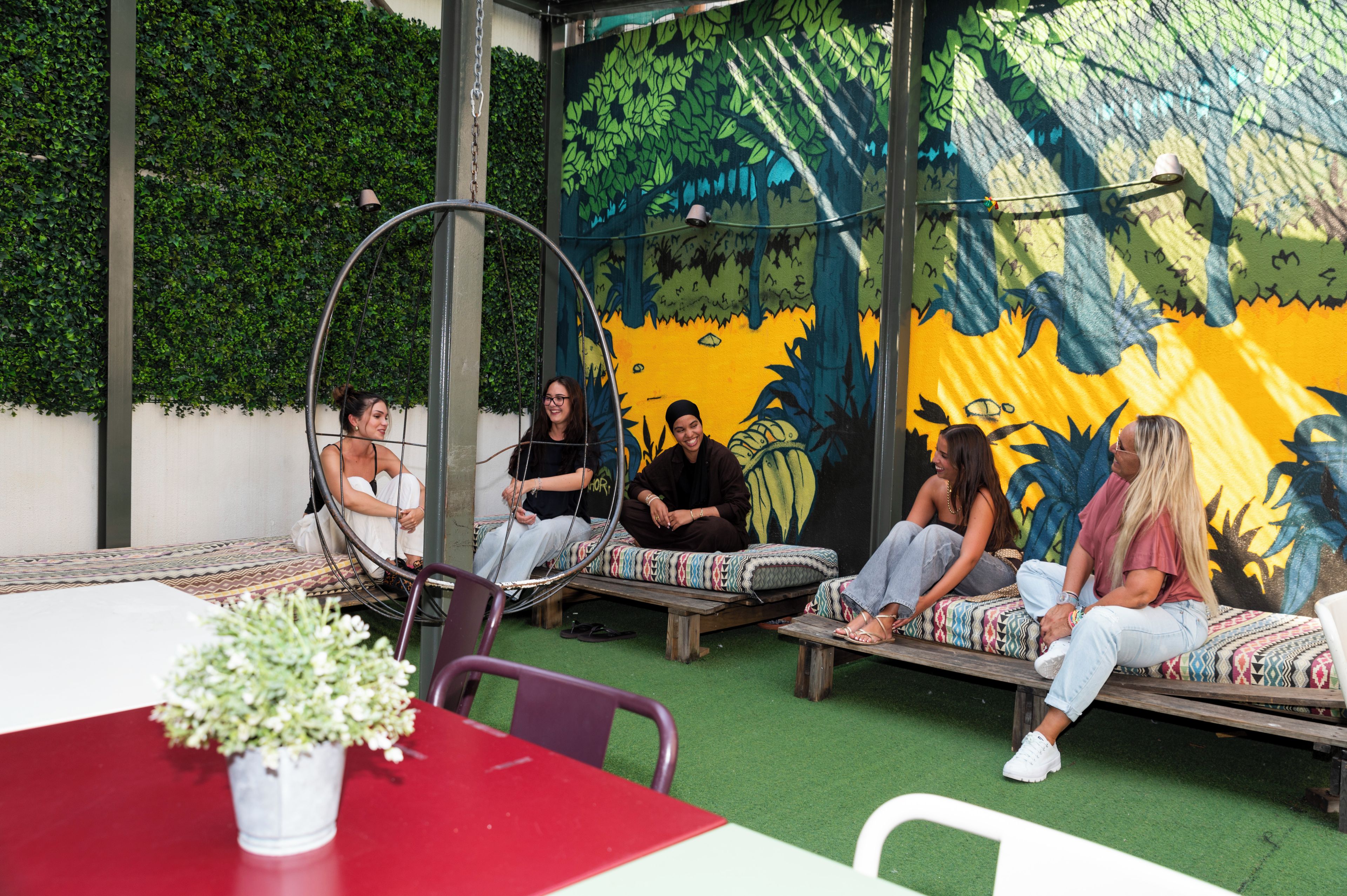 Un grupo de personas sentadas en un banco circular y bancos de madera al aire libre, con un mural de fondo.