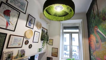 Ein Zimmer mit Deckenleuchte, gerahmten Kunstwerken, einem Fenster mit Blick auf die Straße und einem an der Wand befestigten Regal mit Dekorationen.