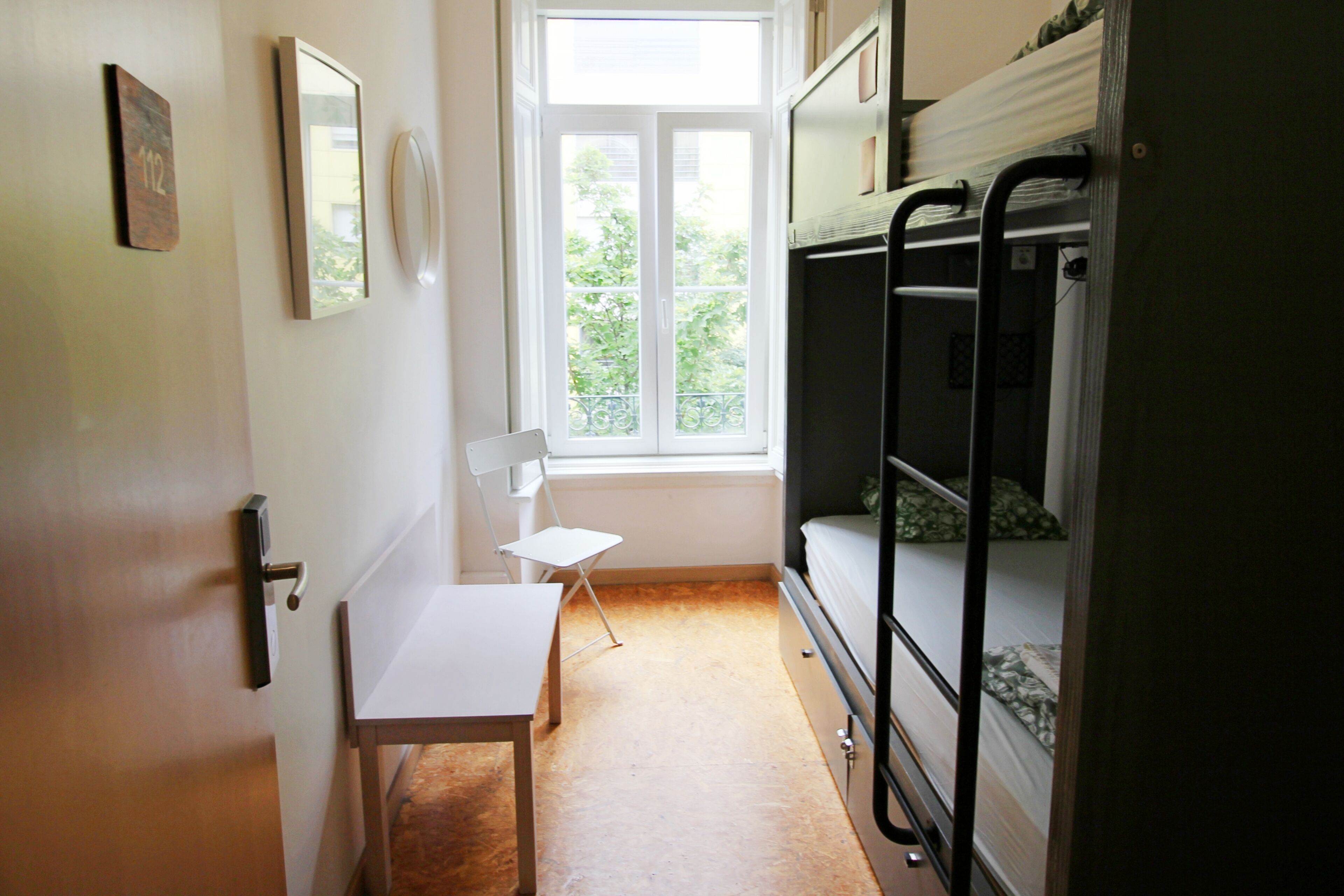 A narrow room with bunk beds, a small table, and a chair near a window.