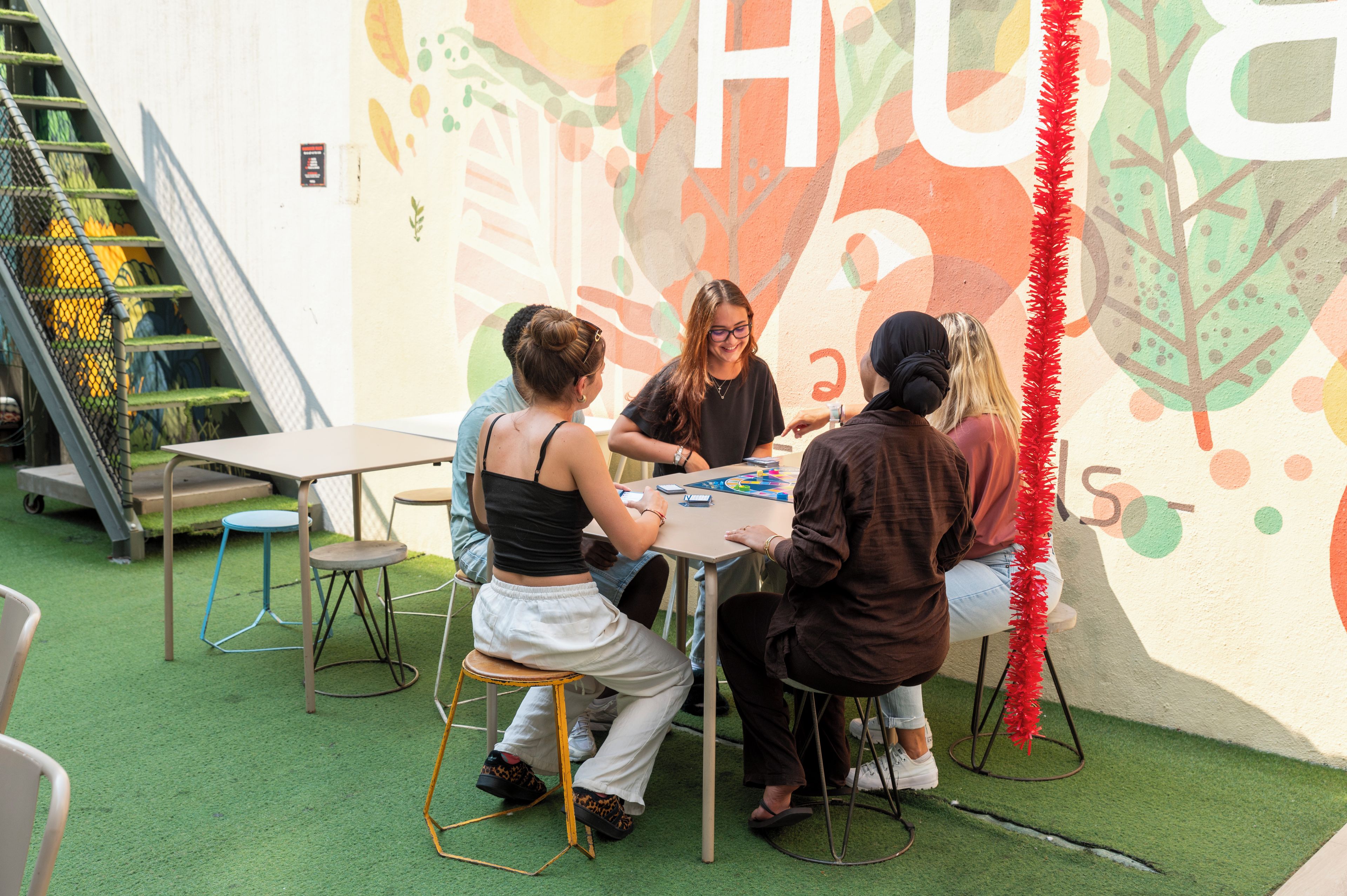 Un grupo de personas sentadas alrededor de una mesa en un espacio al aire libre, con un mural de fondo.