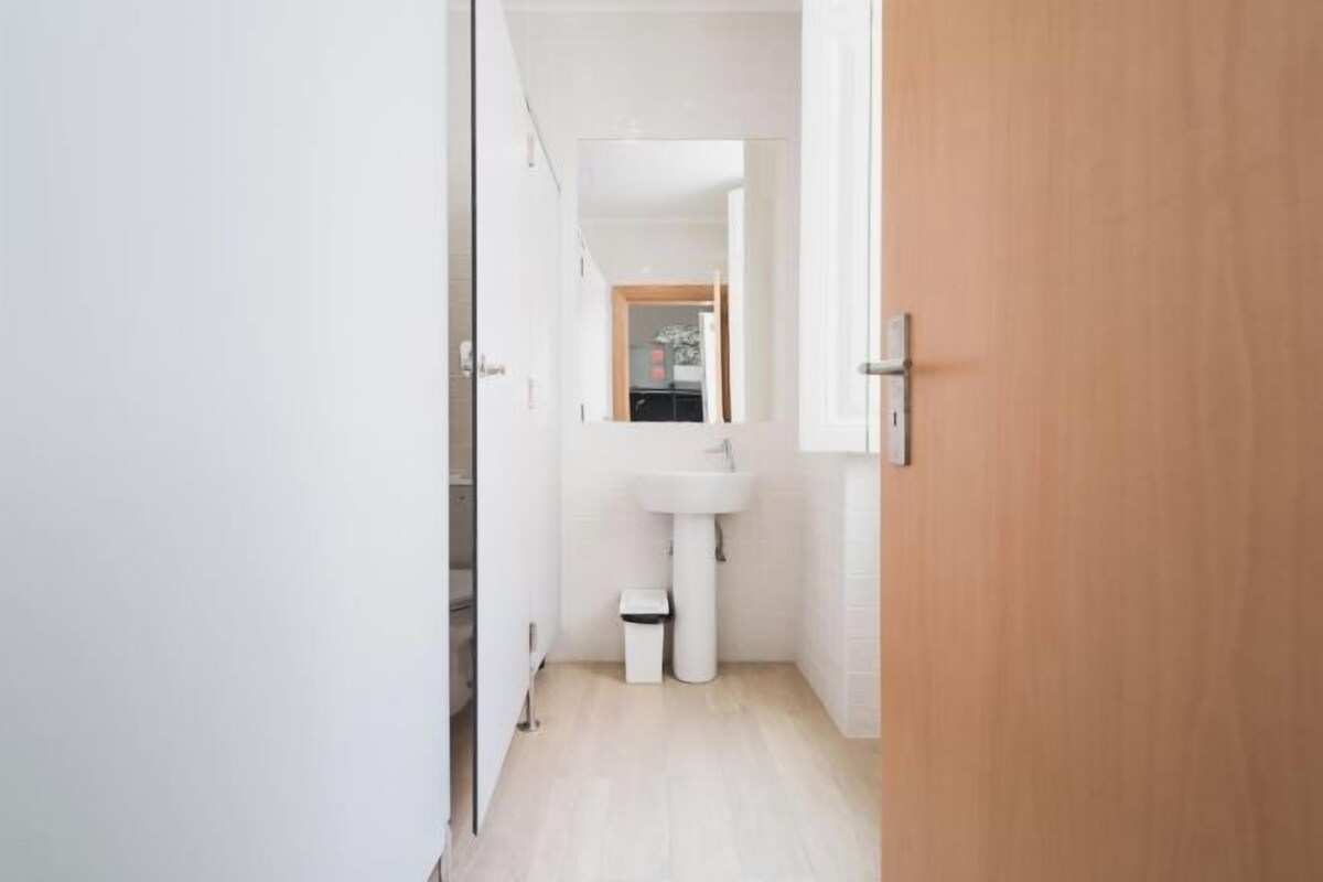 Un pasillo estrecho con un lavamanos blanco y un bote de basura, que da a una puerta de madera.