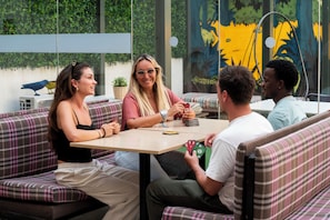 Un groupe de personnes est assis à une table dans un café en plein air, avec vue sur un espace verdoyant et une fresque murale en arrière-plan.