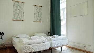 A room with two beds, a desk with a chair, a window with curtains, and wall-mounted artwork.
