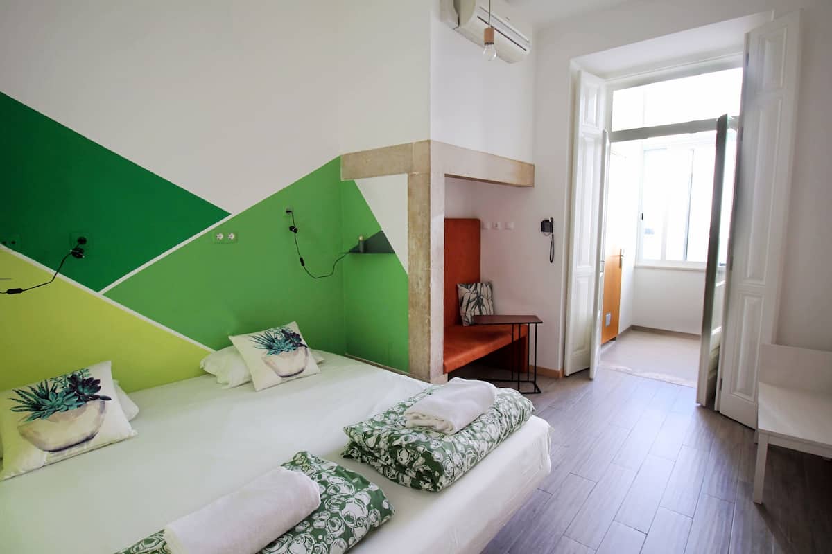Moderni hotellihuone, jossa on sänky, vihreät ja valkoiset seinät, pieni oleskelualue ja näkymä avoimesta ovesta.