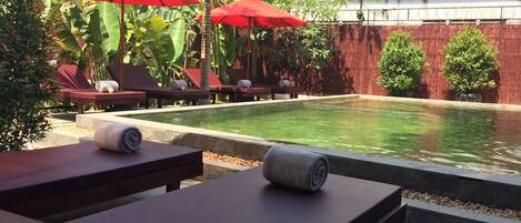 A poolside area with lounge chairs and red umbrellas, surrounded by lush greenery and a brick wall.
