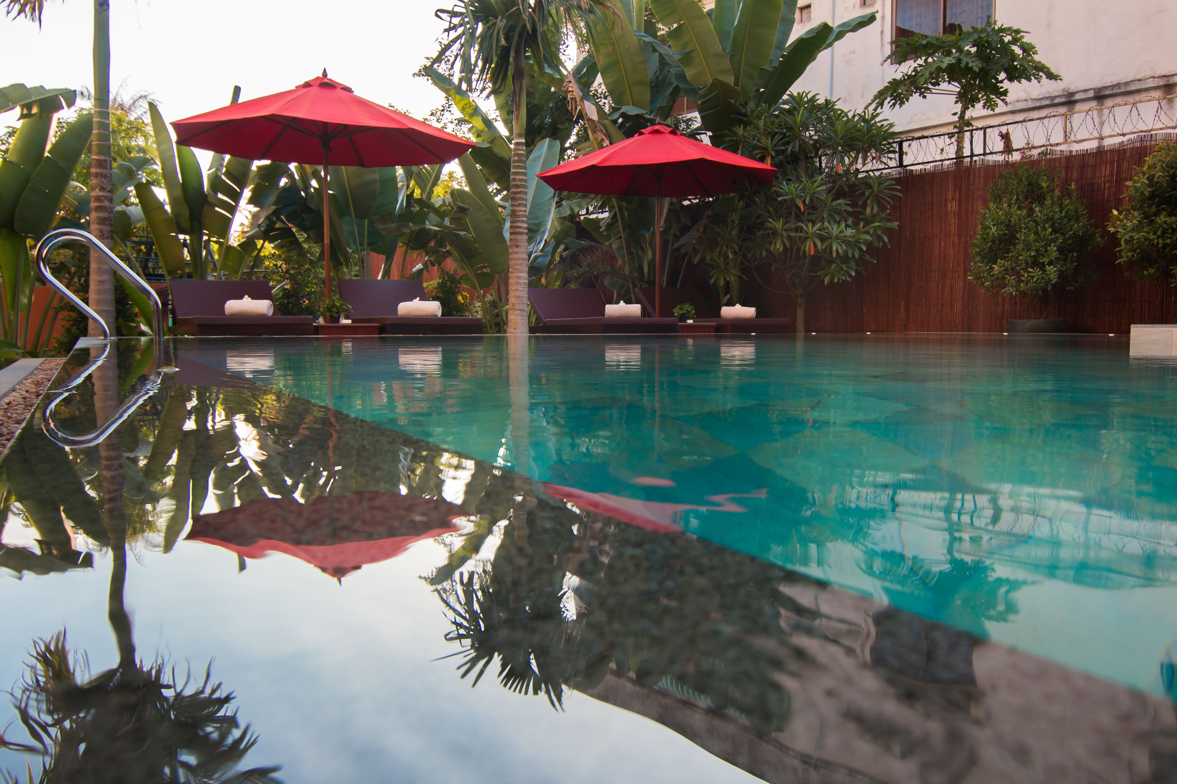 Piscina cristalina com guarda-sóis vermelhos, espreguiçadeiras e plantas tropicais.