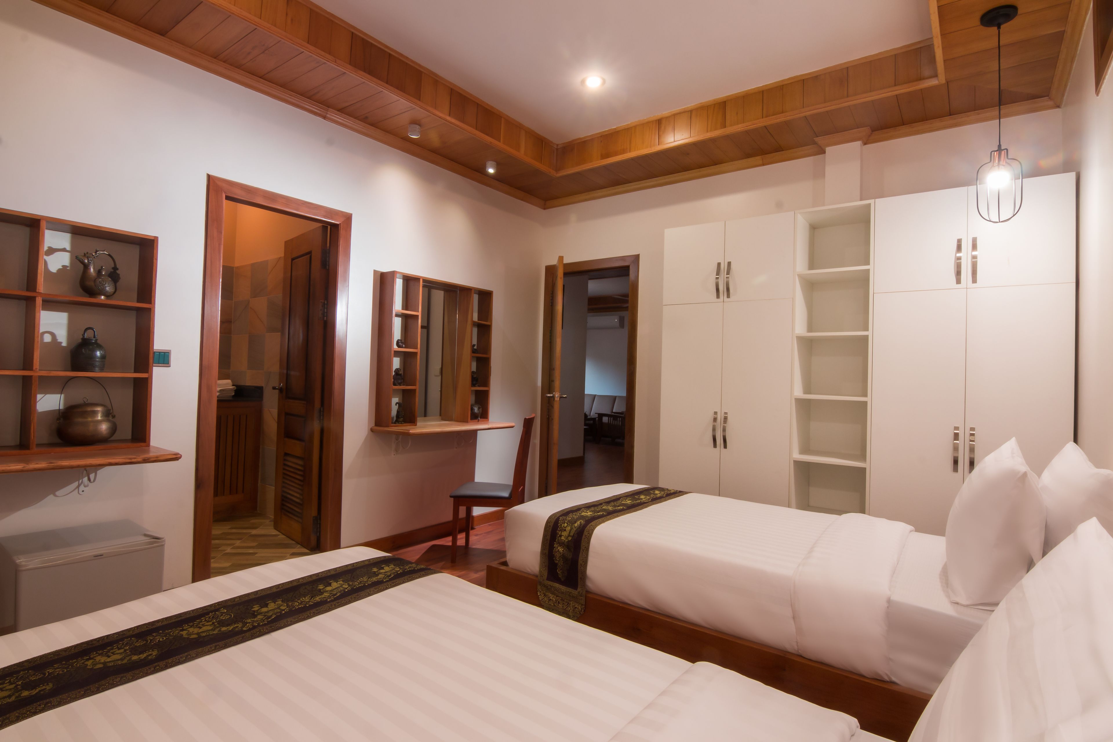 A hotel room with two beds, wooden ceiling, built-in wardrobes, and a small desk with a chair.
