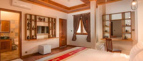 A hotel room with a large bed, wooden ceiling, built-in shelves with decorative items, a small desk, and a window with curtains.