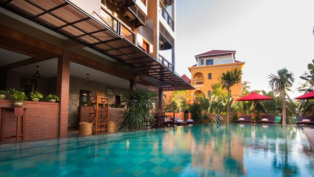 A swimming pool with lounge chairs and umbrellas, surrounded by plants and a building with balconies. - BB Angkor Residence (Siem Reap)