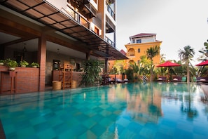 A swimming pool with lounge chairs and umbrellas, surrounded by plants and a building with balconies. - BB Angkor Residence (Siem Reap)