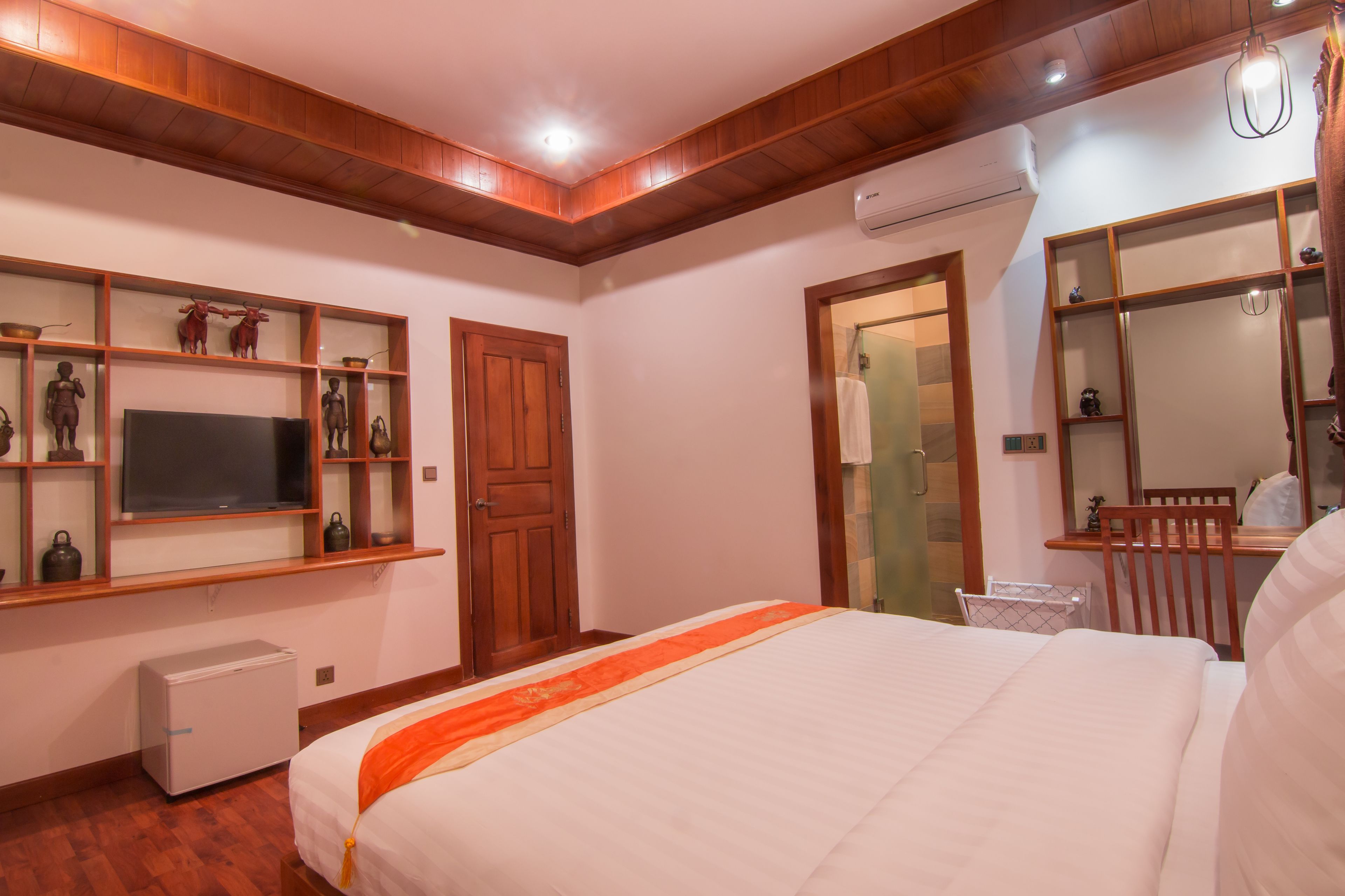 Habitación de hotel con cama, televisión, refrigerador pequeño, estante de madera con objetos decorativos y espejo.