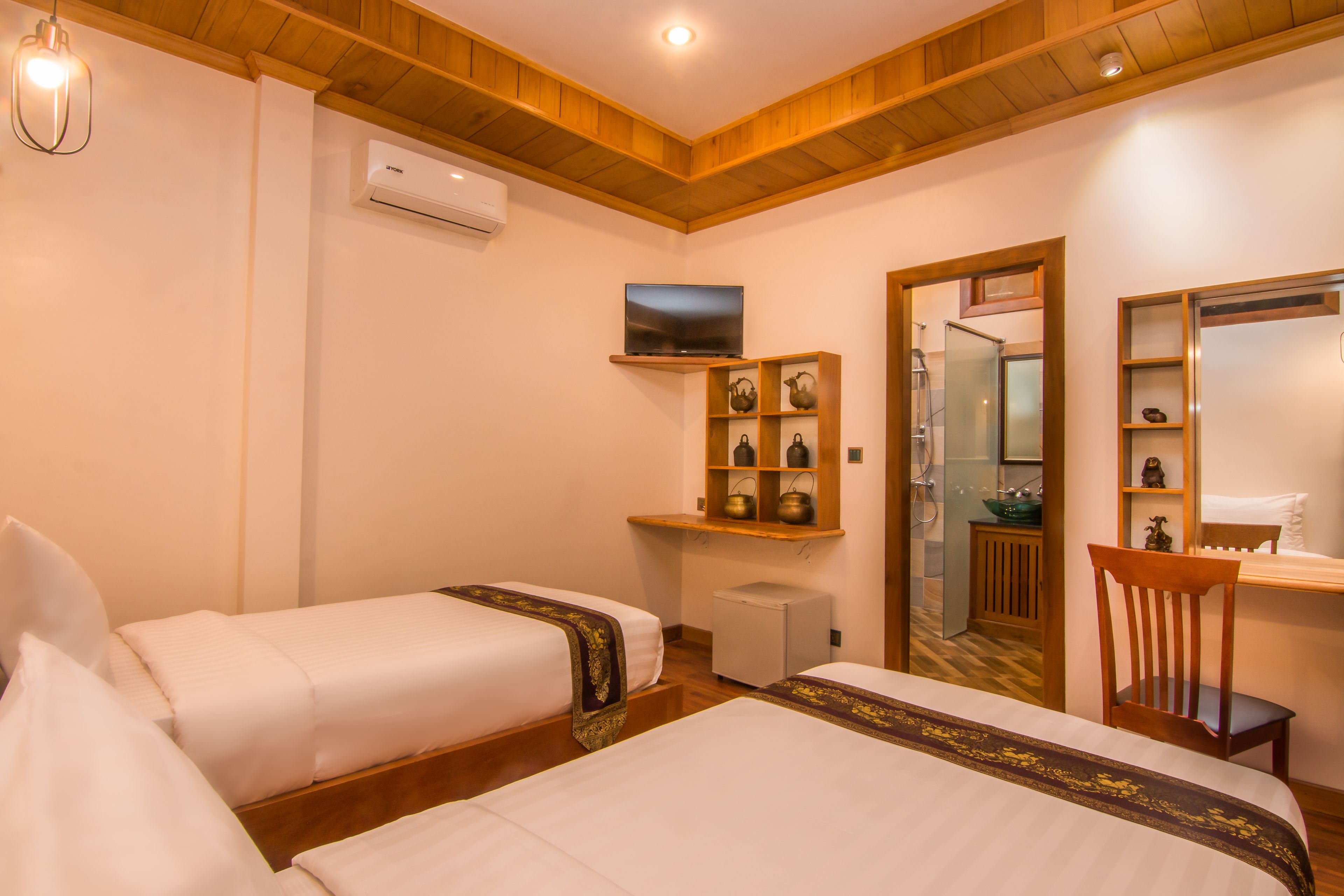 Ein Hotelzimmer mit zwei Betten, einem Fernseher, einer kleinen Kühlbox, einem Spiegel und einem Holzboden mit Dekorationen.