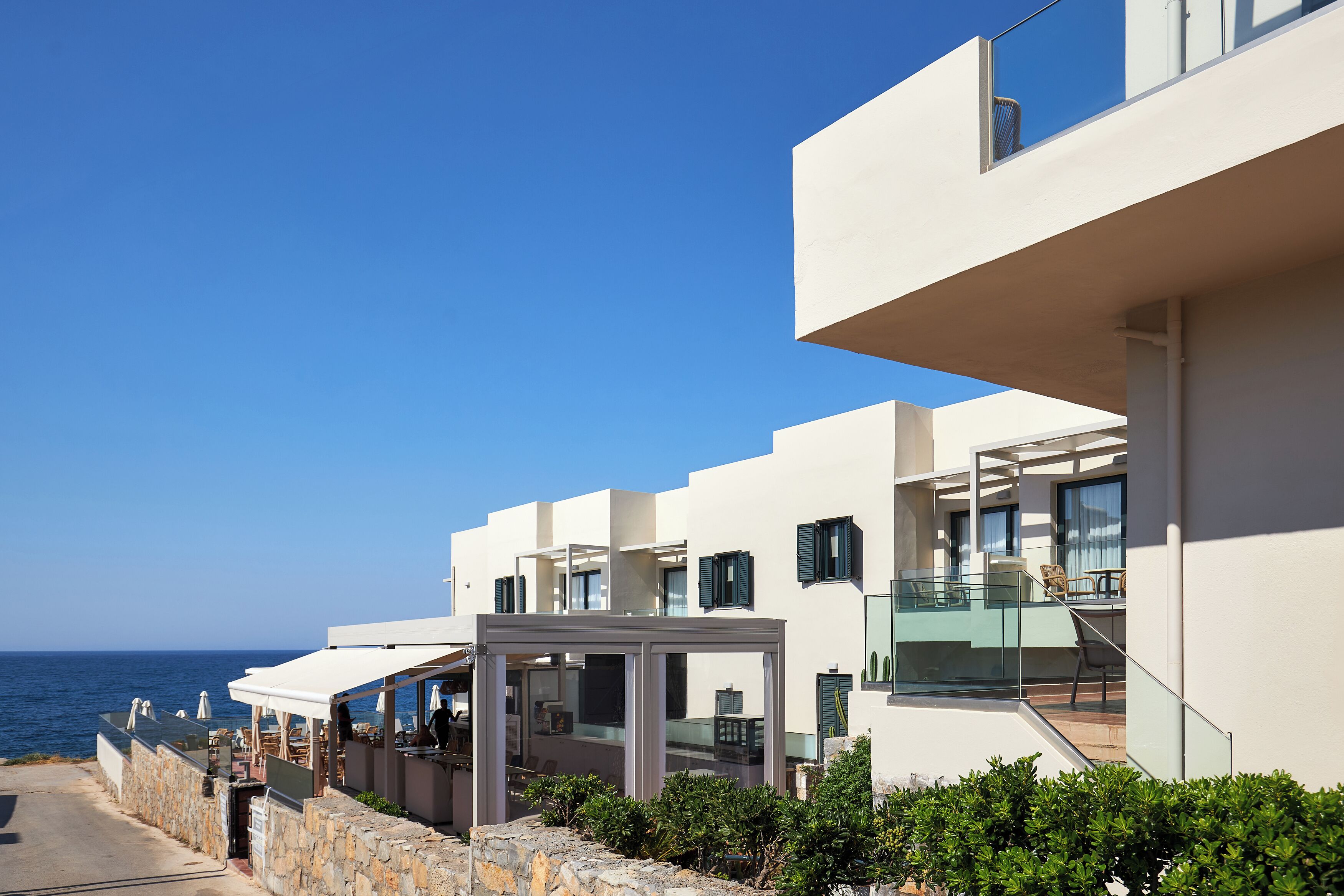 A modern coastal hotel with a terrace, outdoor seating, and a view of the sea.