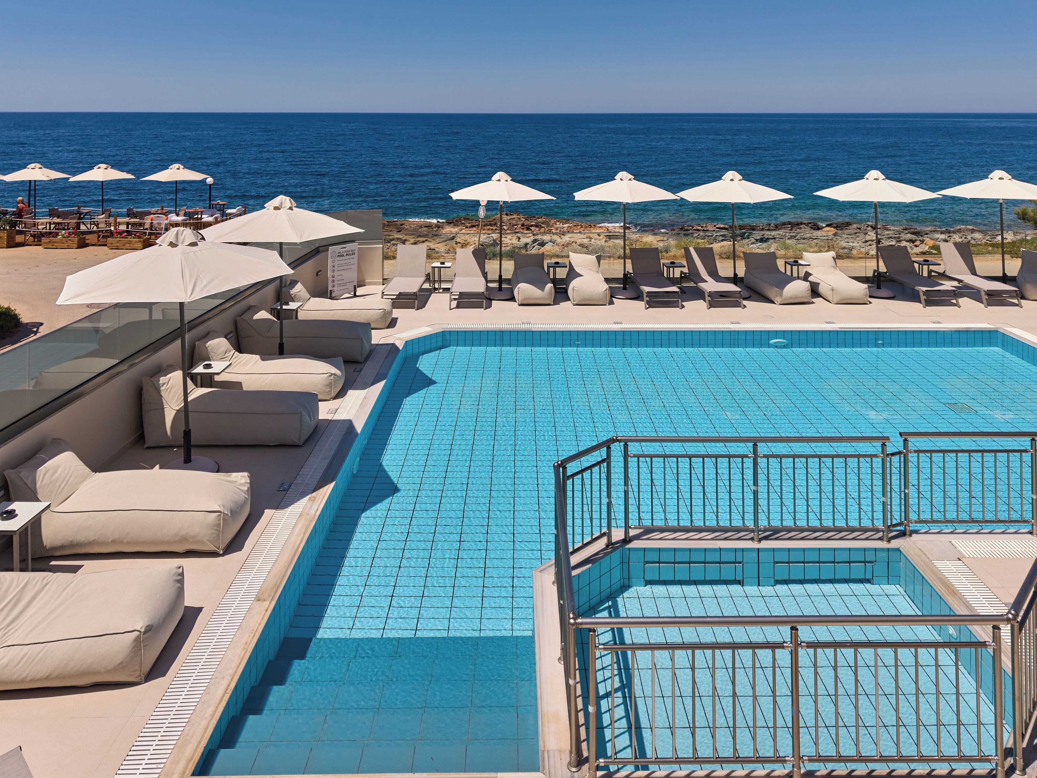 Zona de piscina con sillas de descanso y sombrillas, vista despejada al mar y piscina con azulejos.