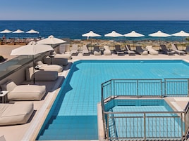 Zona de piscina con sillas de descanso y sombrillas, vista despejada al mar y piscina con azulejos.