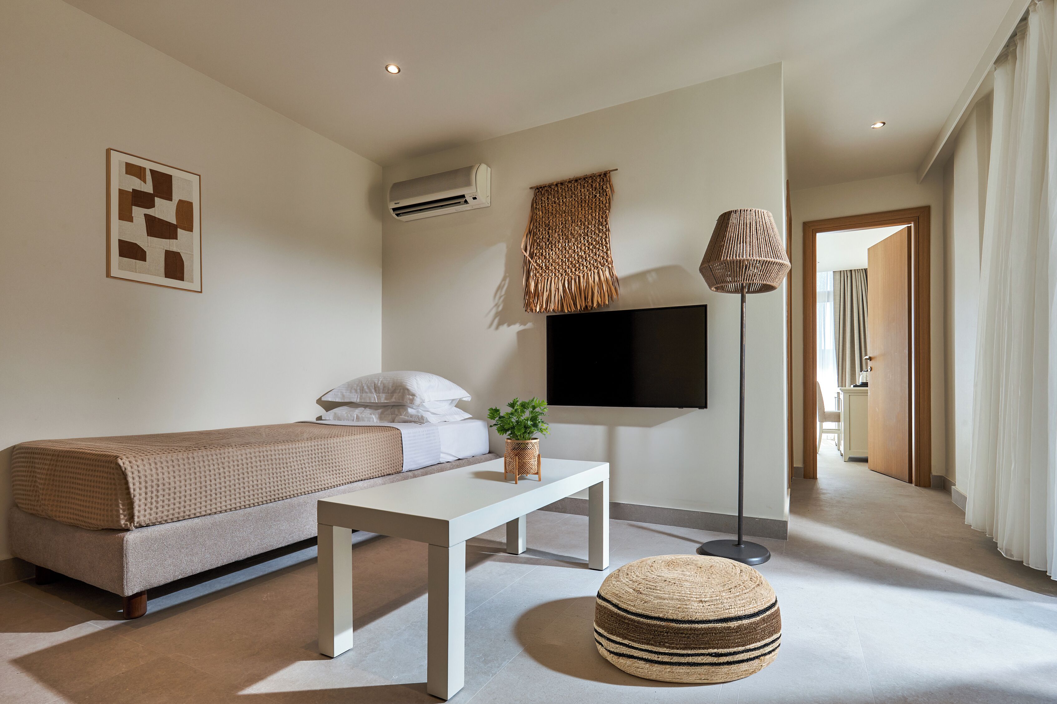 Una habitación de hotel moderna con una cama, un televisor de pantalla plana, una mesita pequeña, una planta en maceta y un otoman tejido.