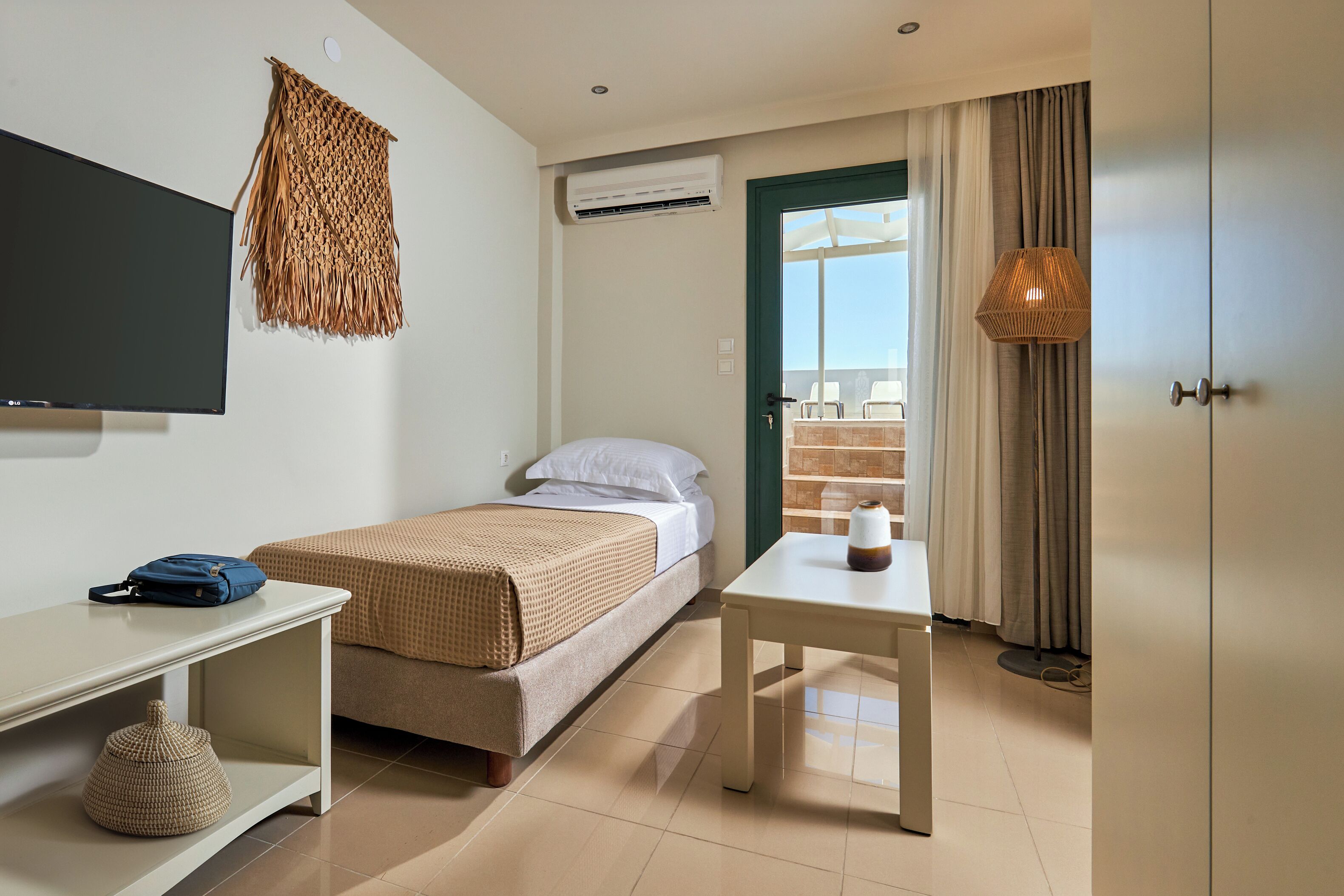 A hotel room with a bed, a desk, a TV, an air conditioner, a window with curtains, and a view of the outside.