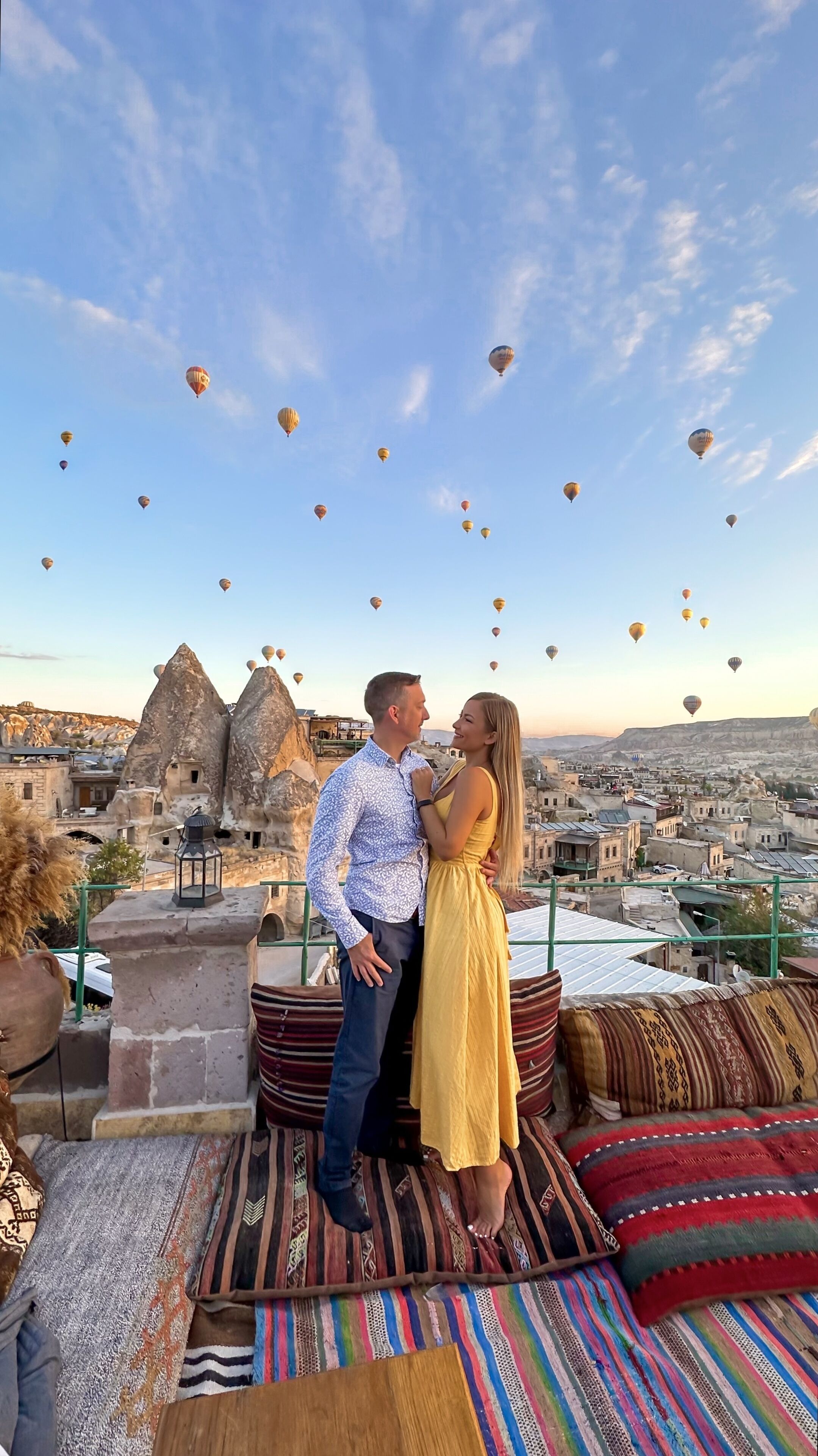 Una pareja parada en una terraza en la azotea, con vistas a un paisaje urbano y globos aerostáticos en el cielo.