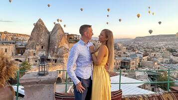 Una pareja parada en una terraza en la azotea, con vistas a un paisaje urbano y globos aerostáticos en el cielo.