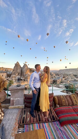 Una pareja parada en una terraza en la azotea, con vistas a un paisaje urbano y globos aerostáticos en el cielo.