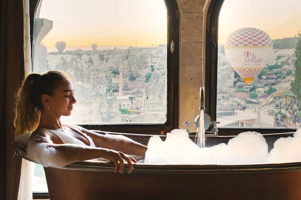 Una mujer relajándose en una bañera con vista a un paisaje urbano a través de ventanas arqueadas.