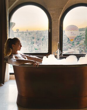Una mujer relajándose en una bañera con vista a un paisaje urbano a través de ventanas arqueadas.