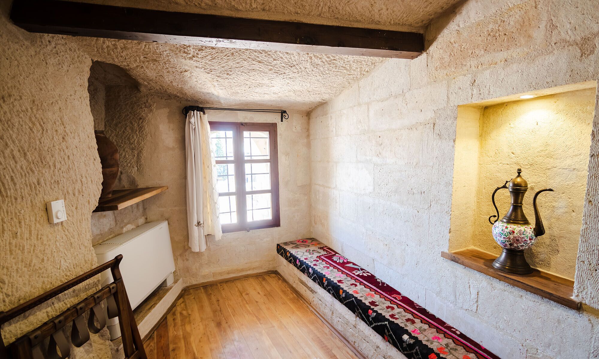 Une petite pièce étroite avec un banc en bois, un radiateur, une fenêtre avec des rideaux et une étagère sur laquelle est posé un objet décoratif.