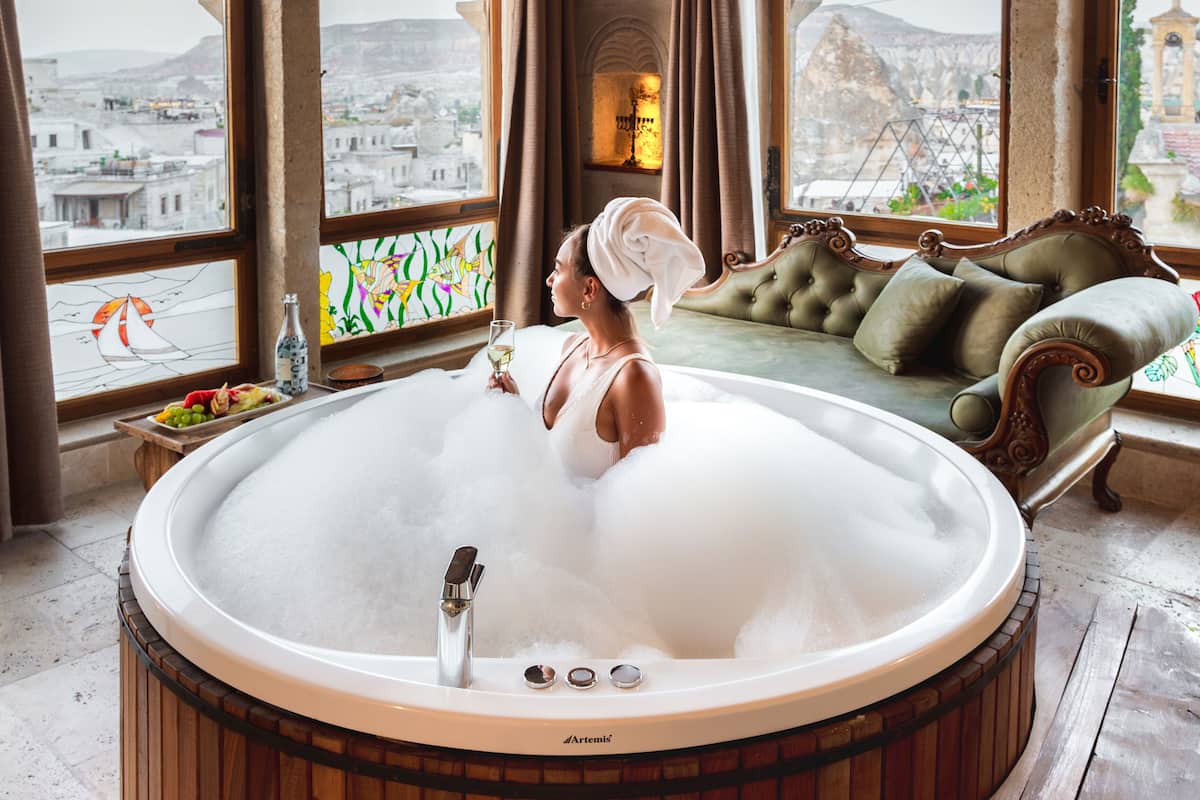 Una mujer en un jacuzzi, con una toalla en la cabeza, suelo de madera, un sofá acolchado y vistas de un paisaje urbano a través de grandes ventanales.