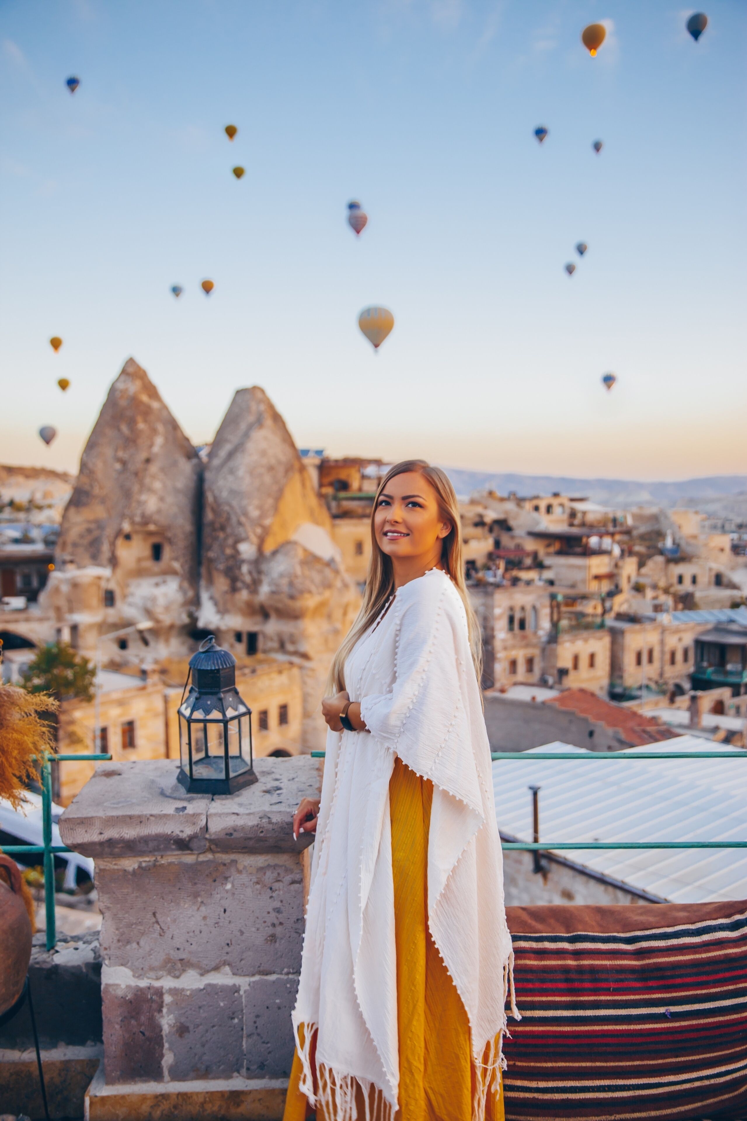 Una persona parada en un balcón con vistas a un paisaje urbano y globos aerostáticos en el cielo.