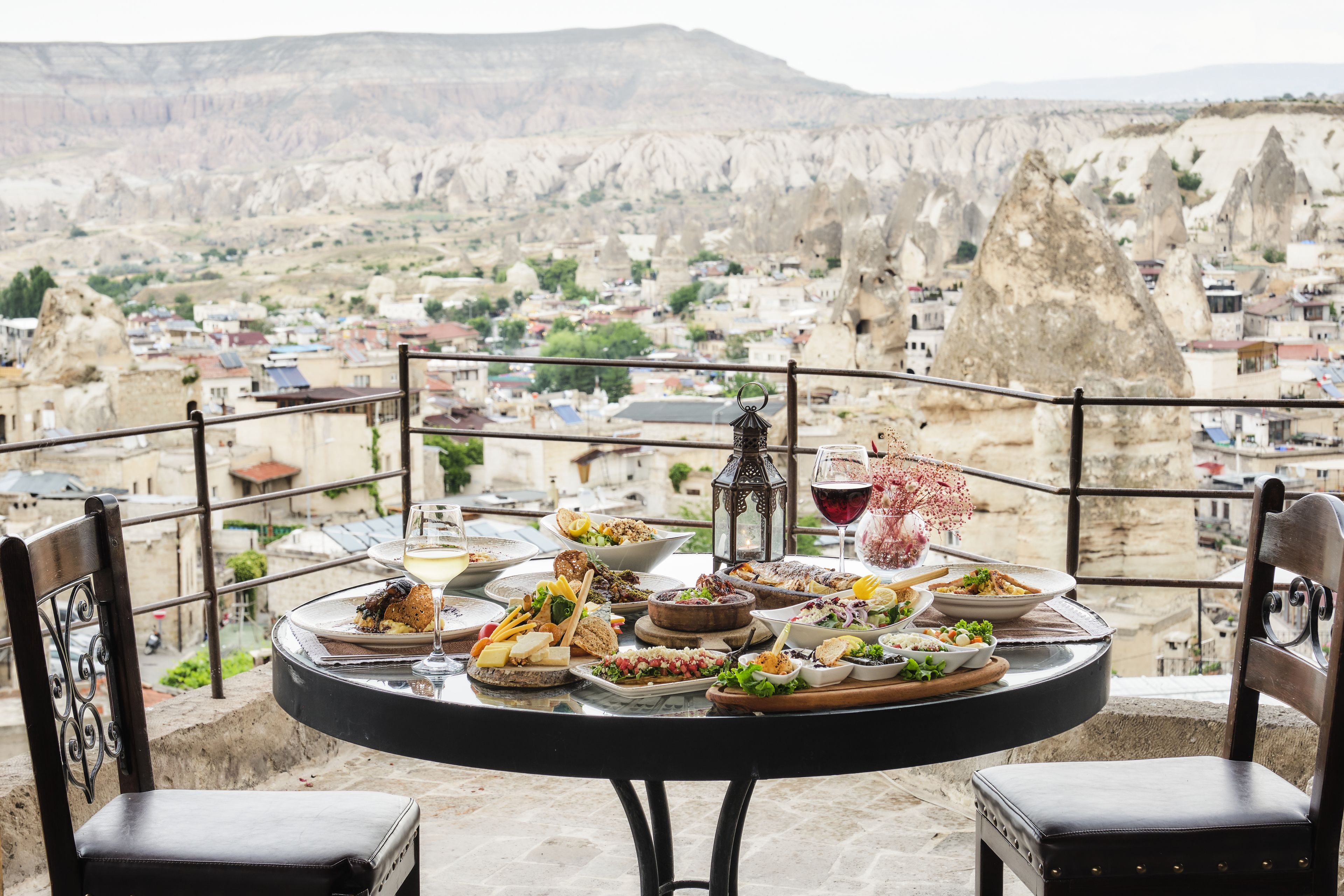 A table set with various dishes and a wine glass, overlooking a town with distinctive rock formations.