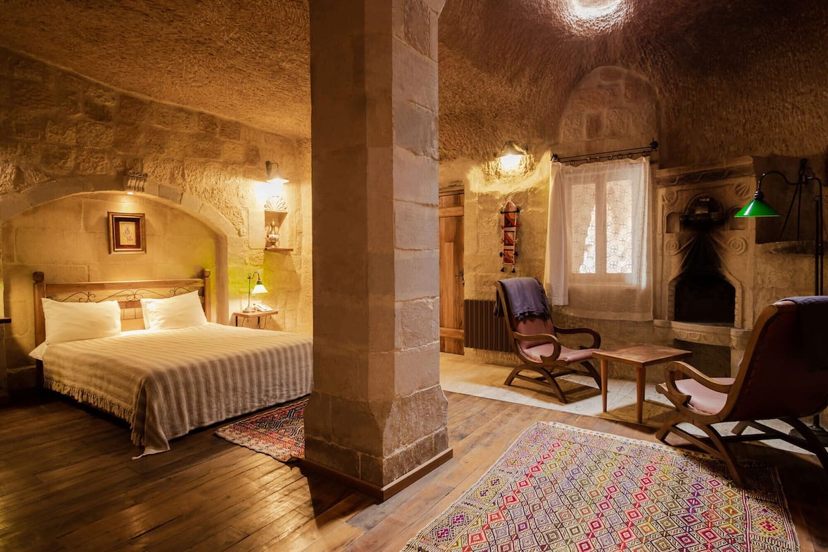 Una habitación rústica con una cama, una silla de madera, una chimenea y un muro de piedra.