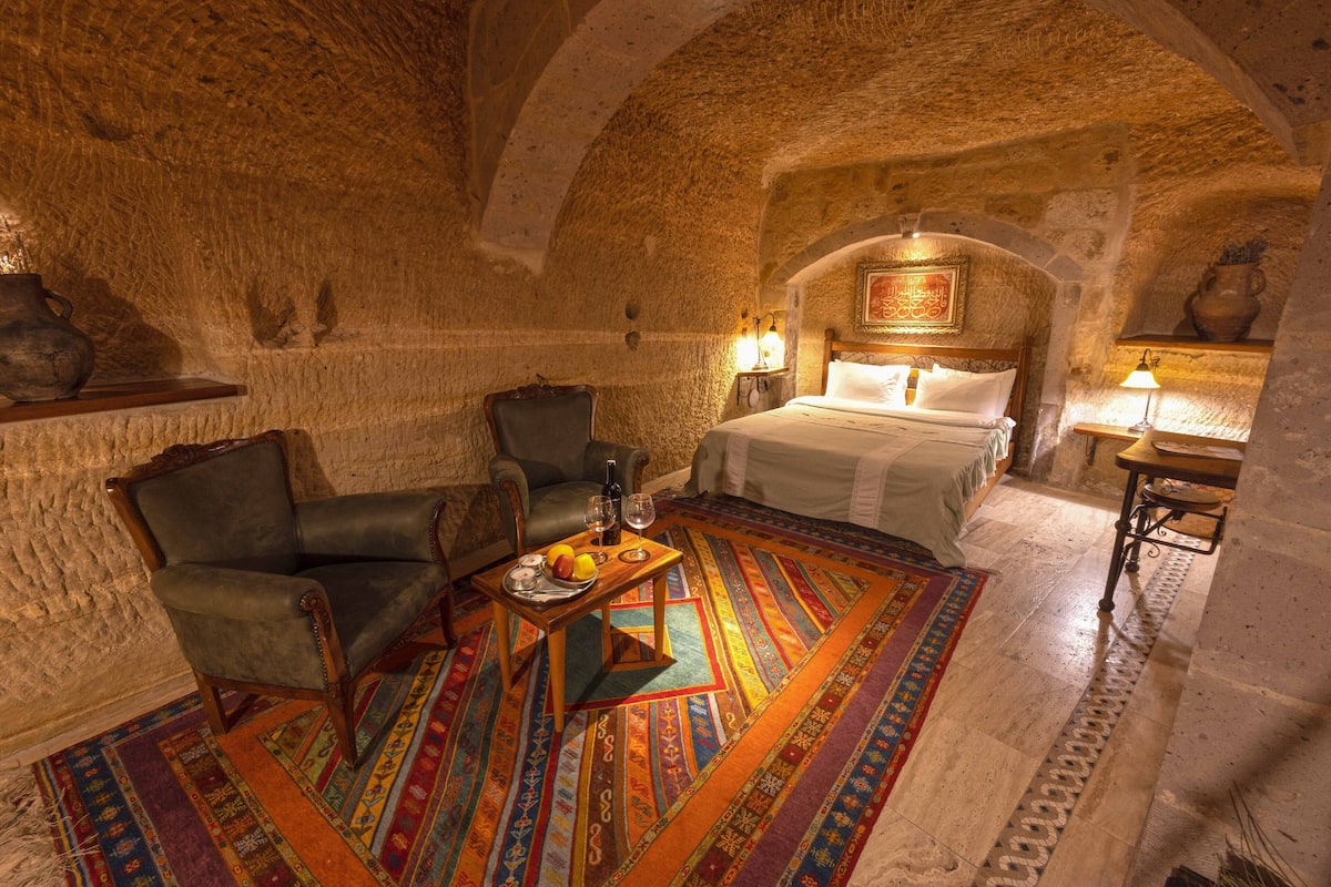 Una habitación con pared de piedra, una cama con sábanas blancas, una silla de madera, una mesa con frutero y una alfombra colorida.