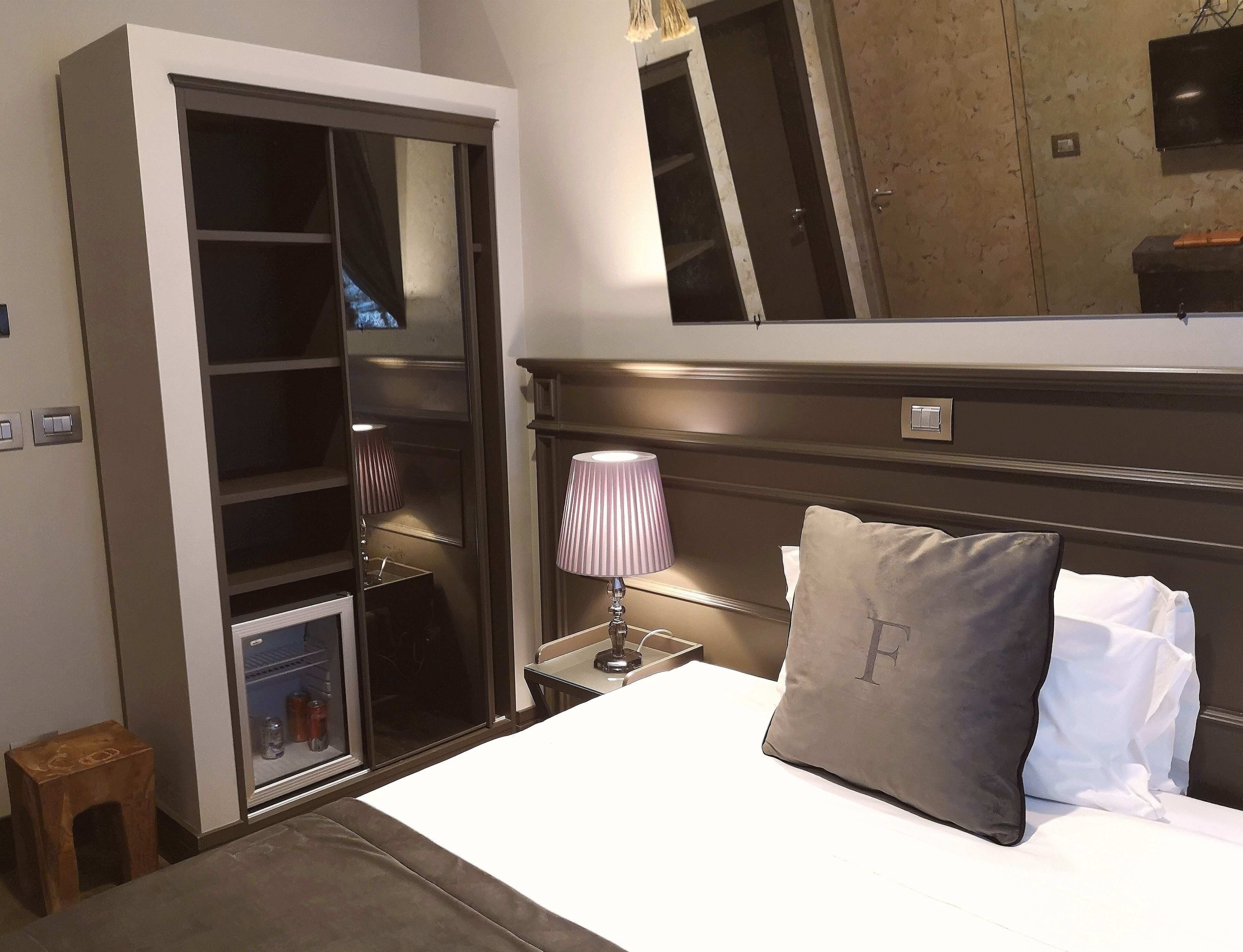 Un dormitorio con cama, mesita de noche con lámpara, armario y espejo en la pared.