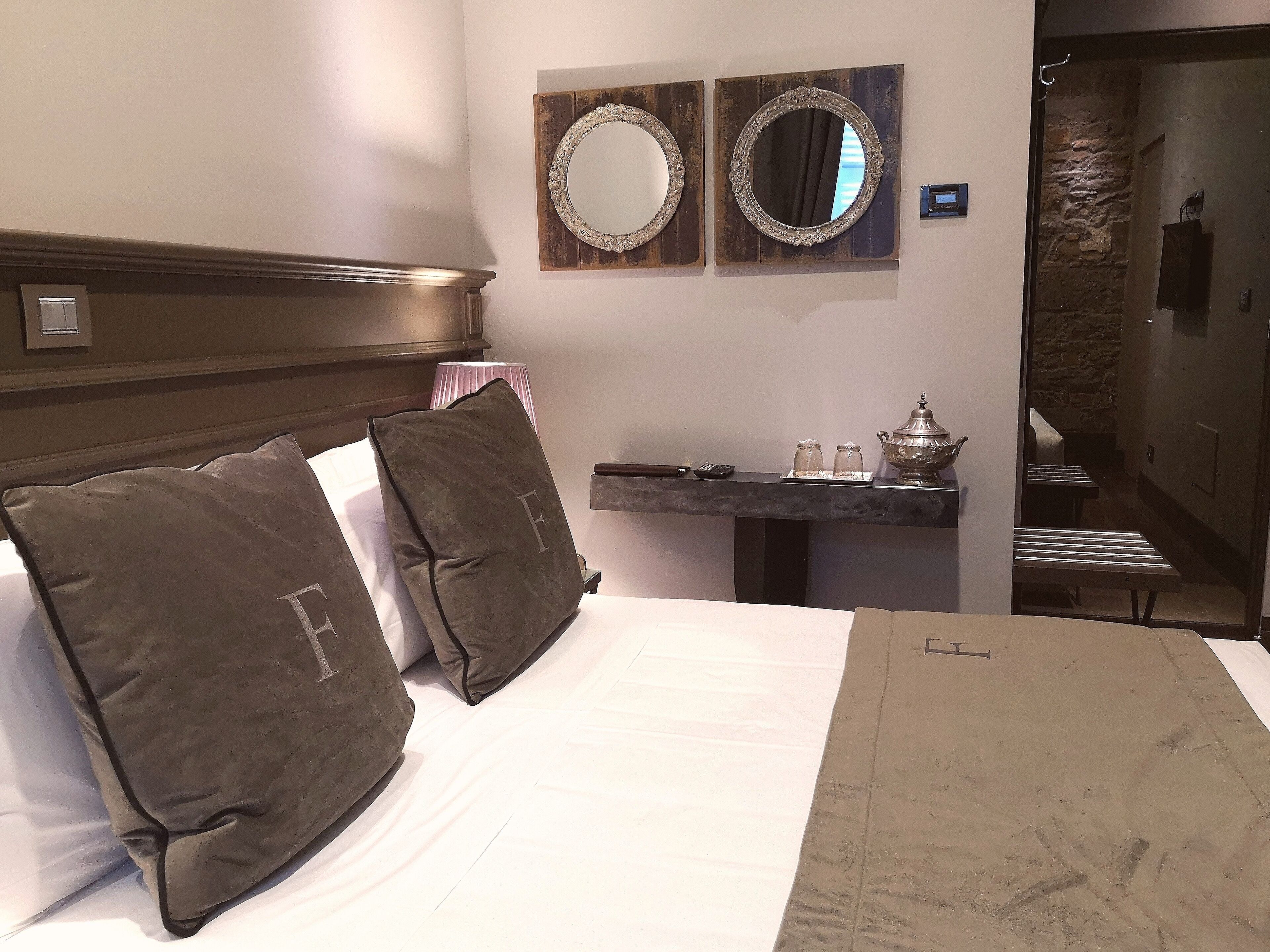 Een hotelkamer met een bed, twee kussens, een nachtkastje met een lamp, een spiegel en een klein tafeltje met een theepot.