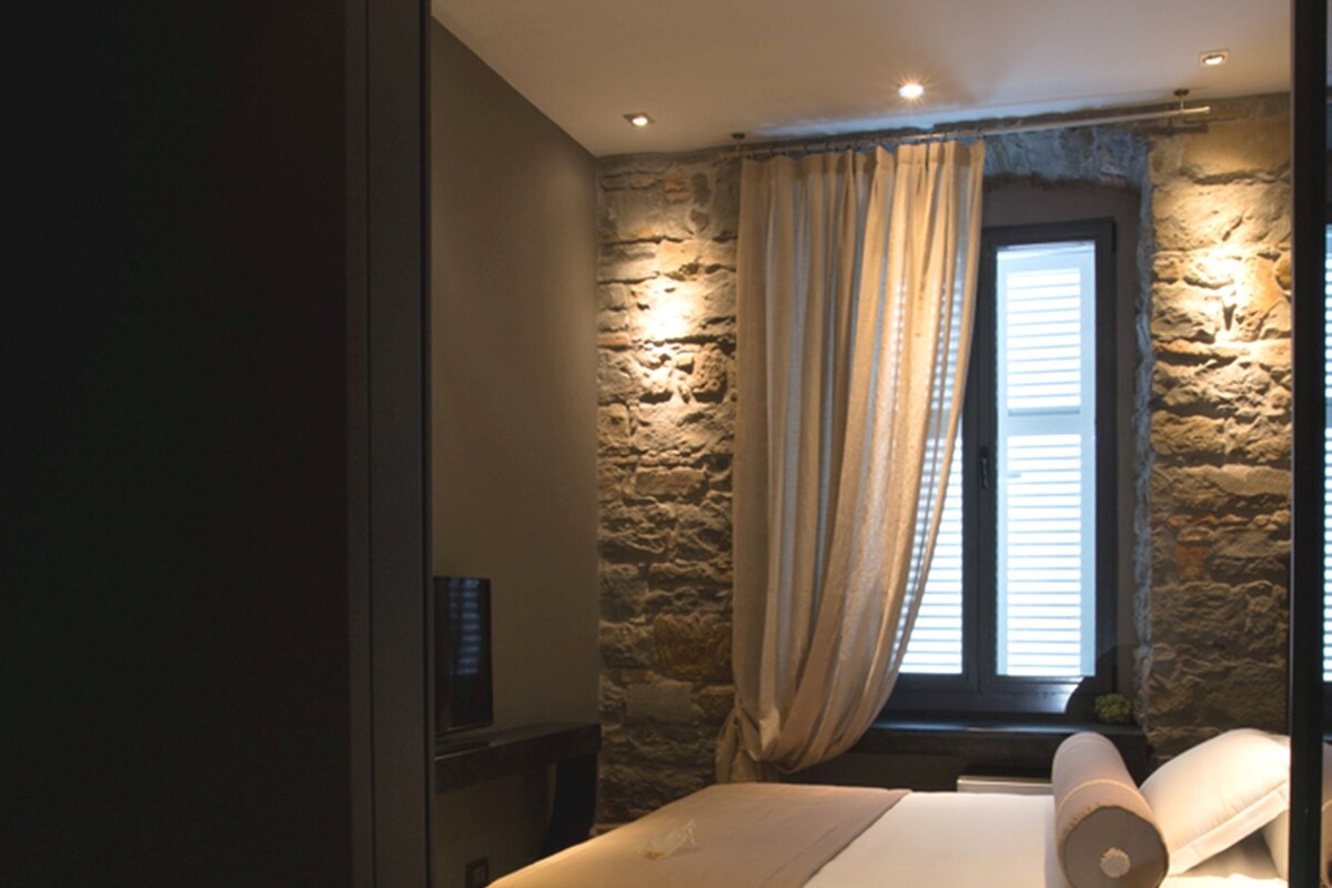 A bedroom with a stone wall, a wooden floor, a bed with a beige blanket, and a window with curtains.