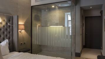 A modern hotel room with a tufted headboard, a bedside lamp, a glass shower enclosure, and a dark door.